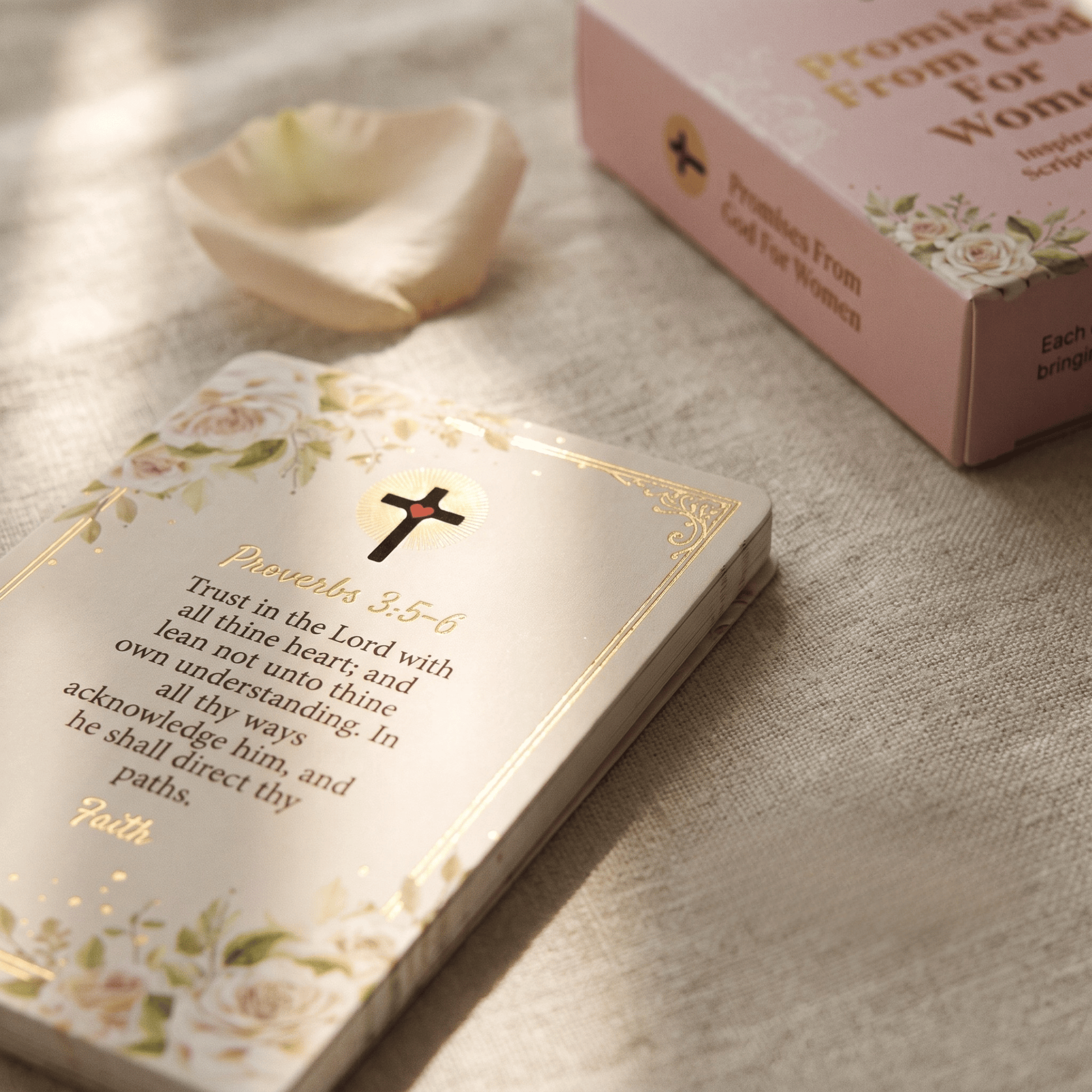 A close-up of a small floral prayer card with a black cross and Bible verse from Proverbs 3:5-6, placed on a textured beige fabric. A matching pink box reading 'Promise From God For Women' sits in the background with a soft rose petal nearby.
